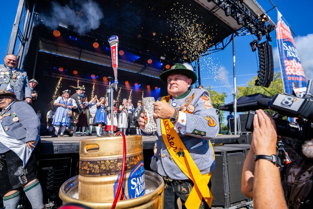 A person in oktoberfest garb with a keg in front of them and people on a stage behind
