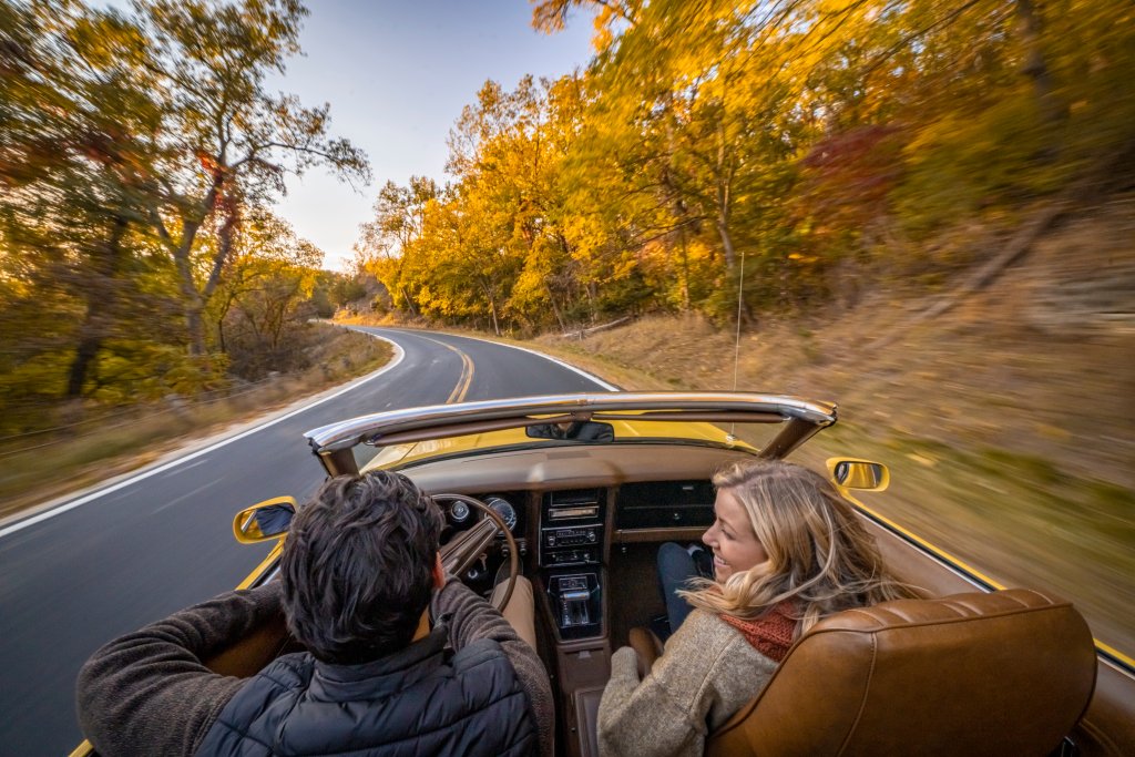 A couple smile as they drive a convertible vehicle with the top down on a paved road during the fall season in beautiful La Crosse, Wisconsin.